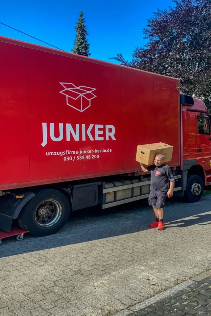 Umzugshelfer der Umzugsfirma Junker trägt einen Karton vor einem roten Umzugs-Lkw mit Firmenlogo.