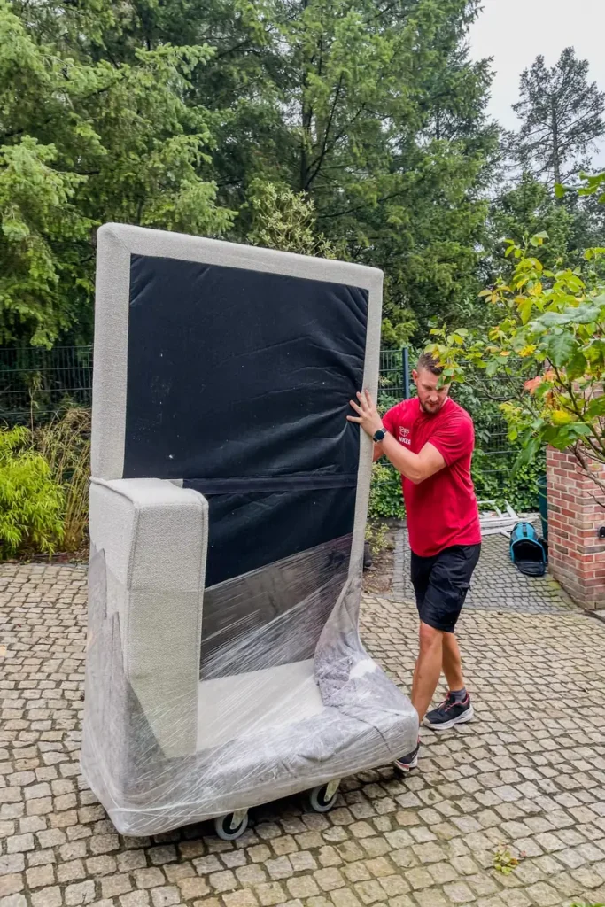 Umzugshelfer der Umzugsfirma Junker transportiert verpacktes Möbelstück auf Rollwagen.