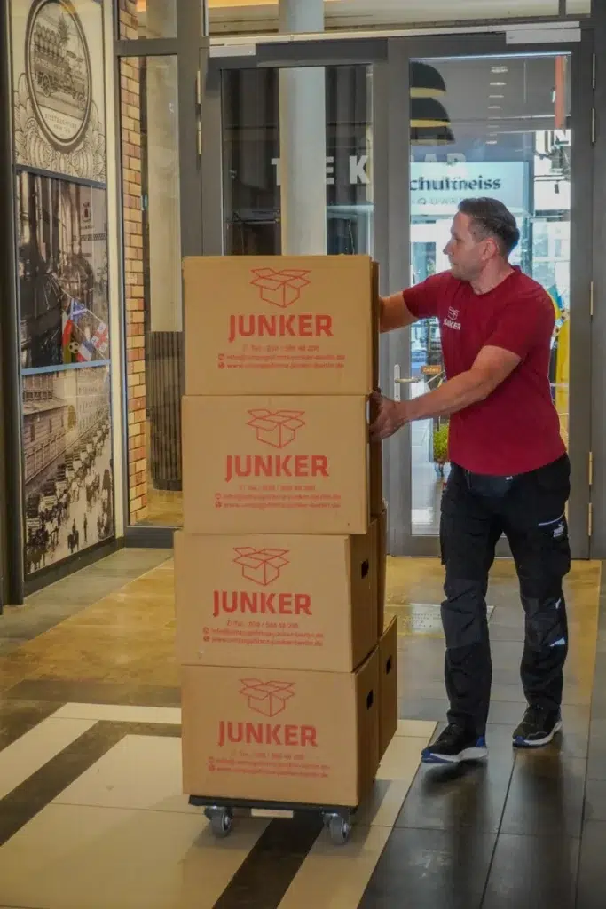 Junker-Mitarbeiter transportiert gestapelte Umzugskartons auf einem Rollwagen im Eingangsbereich